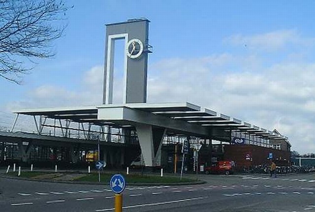 station Almelo Opvallende elementen zijn het grote dak van het stationsgebouw met overstek aan het stationsplein en de klokkentoren. Het dak wordt gedragen door een brede hoge balk die aan stationspleinzijde rust op een V-vormige kolom. De stationshal heeft glazen gevels. Aan beide zijden van de hal staan, evenwijdig met het spoor, twee lage donkerrode bakstenen volumes.