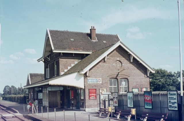 station Beesd Het station gezien vanaf de spoorzijde. Het is goed weer, want de deur naar Post T staat open. Let op het overpad naar het perron, dat toen d.m.v. een simpele ketting beveiligd werd. Links naast het hoofdgebouw zie je het bijgebouw, wat o.a. de toiletten herbergde. Ook leuk: de reclame-affiches van toen.
<P>
Dia: F.A.H. van den Hurk, scan: Frans van den Hurk