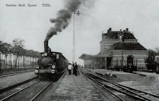 station Tiel Deze op 29 juli 1911 door de post afgestempelde, helaas wat gevlekte ansichtkaart, werd vervaardigd door de Tielse uitgever en drukker D. Mijs. Een sprekender beeld van het stoomtijdperk is haast niet denkbaar, met een rookuitpuffende schoorsteen van de stoomlocomotief, die met de houten personenwagons gereed staat om richting Utrecht, via Culemborg en Geldermalsen te vertrekken. Links is goed te zien, dat de bebouwing aan de kant van de toenmalige gemeente Wadenoyen-Drumpt, nog nagenoeg onbebouwd was. De huizen op de achtergrond zijn die van de Lingedijk. Het stationsgebouw rechts heeft nog zijn 19e eeuwse uiterlijk en de rails voor het goederen/rangeerstation liepen door tot vlak bij de overkapping links op de kaart. De titel van de ansichtkaart ´Station Hollands Spoor´ geeft de naam weer van de spoorwegmaatschappij, die deze lijn beheerde. Pas vele jaren later is de huidige NS door verschillende fusies van spoorwegexploitatiemaatschappijen ontstaan.